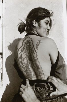 a woman with a tattoo on her back standing next to a wall and looking at the camera