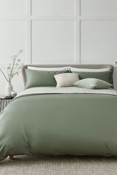 a bed with green sheets and pillows in a white room next to a vase filled with flowers