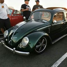 two men standing next to an old green car in a parking lot with other cars