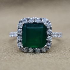 an emerald colored stone surrounded by white diamonds in a halo setting on a sandy surface