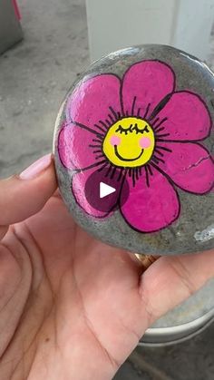 a hand holding a painted rock with a smiley face on it and a pink flower