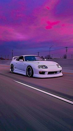 a white sports car driving down the road at dusk with pink clouds in the background