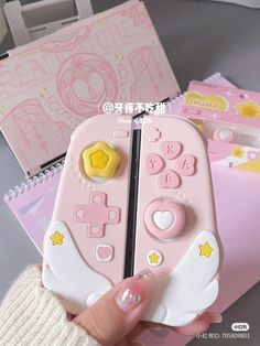 a hand holding a pink and white game controller with yellow stars on the side, in front of some other items