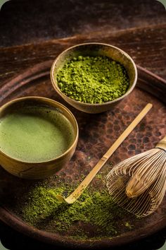 two bowls filled with green powder and whisk on top of a wooden tray