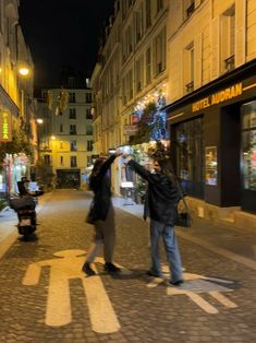 a man and woman standing on the side of a road next to buildings at night