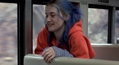 a woman with blue hair sitting on a bus looking out the window and smiling at something