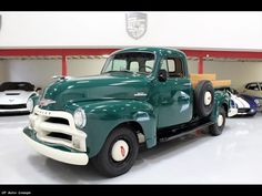 an old green truck is parked in a garage with other cars behind it and on the floor