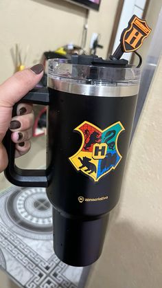 a person holding a black coffee cup with the hogwarts crest and harry potter symbols on it