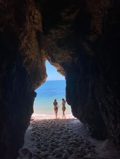 two people standing in the middle of a cave on a beach with blue water behind them