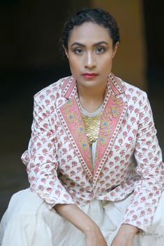 a woman sitting down wearing a white and pink suit with gold accents on her lapel