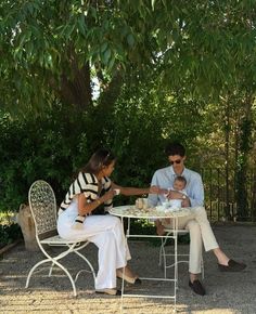 two people sitting at a table eating food and drinking tea in the shade of a tree