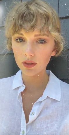 a woman with blonde hair wearing a white shirt and looking at the camera while standing in front of a brick wall