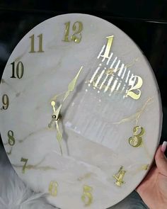 a white clock with gold numbers on the face is being held by a woman's hand