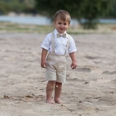 a young boy wearing a bow tie and suspenders standing in the sand at the beach