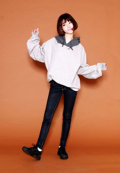 a woman standing in front of an orange wall wearing black pants and a gray sweatshirt