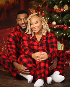 a man and woman dressed in red and black plaid pajamas sitting next to a christmas tree