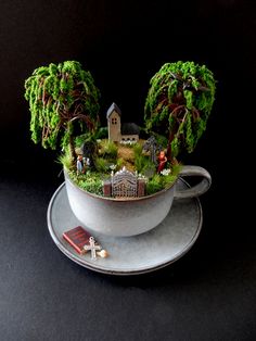 a cup filled with miniature trees and plants on top of a saucer next to a book