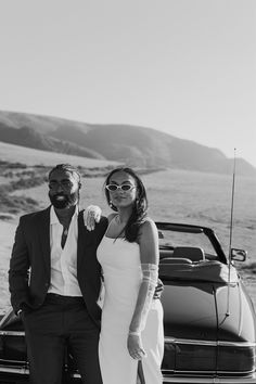 a man and woman standing next to each other in front of a car on the beach