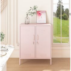 a pink cabinet sitting in front of a window next to a vase with a plant