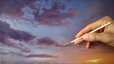 a hand holding a pencil in front of a painting with purple and blue clouds behind it