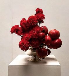 a silver vase filled with red flowers and apples on top of a white pedestal in front of a wall