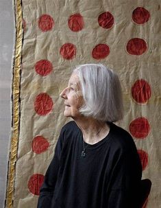 an older woman is sitting in front of a wall with red circles on it and she has white hair