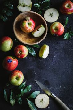 some apples are in a wooden bowl on a black surface with leaves and a knife