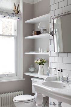 a white toilet sitting next to a sink in a bathroom under a window with shelves above it