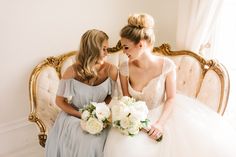 two bridesmaids sitting on a couch with bouquets in their hands and looking at each other