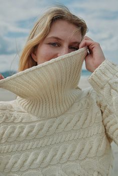 a woman wearing a white sweater and holding her hand to her face while standing on the beach