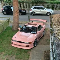 a pink car parked next to a tree on the side of a road in front of a fence