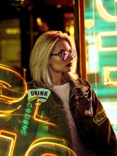 a blonde woman wearing sunglasses and a jacket with the word drink on it is standing in front of neon lights