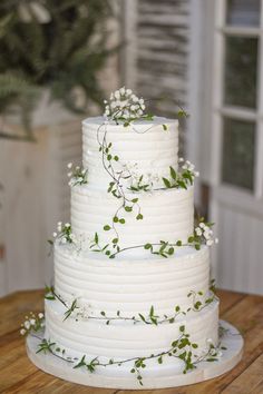 a three tiered cake with white flowers and greenery on the top is sitting on a wooden table