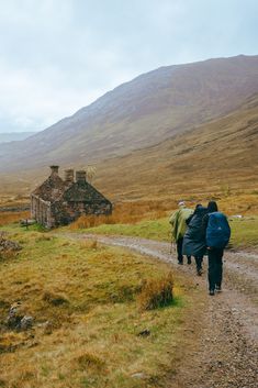 two people walking down a dirt road towards an old stone house in the middle of nowhere