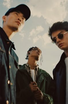 three young men standing next to each other under a cloudy blue sky with clouds in the background