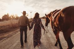 a man and woman walking down a dirt road holding hands with a horse in the background