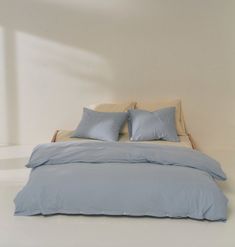 a bed with blue sheets and pillows in a white walled room, next to a window