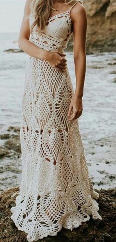 a woman wearing a white crochet dress standing on rocks near the ocean with her arms around her waist