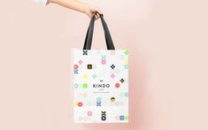 a hand holding a white shopping bag with the words kindo printed on it against a pink background