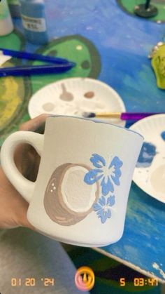 a person holding a coffee cup in front of paintbrushes and paper plates on a table