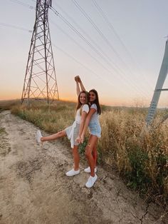 two girls are standing in the middle of a dirt road with power lines behind them