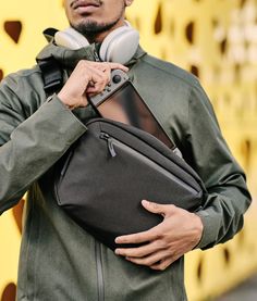 a man with headphones is holding his bag and looking off to the side as he stands in front of a yellow wall