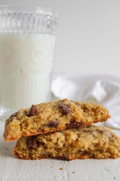 two oatmeal chocolate chip cookies next to a glass of milk on a white table