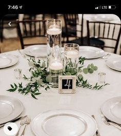 the table is set with white plates and silverware, candles and greenery on it