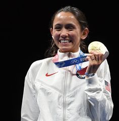 a woman holding up a gold medal in her right hand and wearing a white jacket