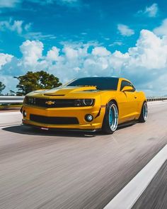 a yellow chevrolet camaro driving down the road with blue sky and clouds in the background