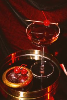 a red drink in a glass on top of a metal tray with two cherries