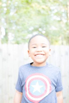 a young boy wearing a captain america t - shirt smiles at the camera while standing in front of a fence