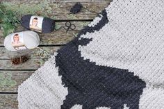 a crocheted cow blanket next to yarn and scissors on a wooden table with pine cones