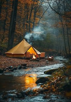 a tent set up next to a stream in the woods with fire and chairs around it